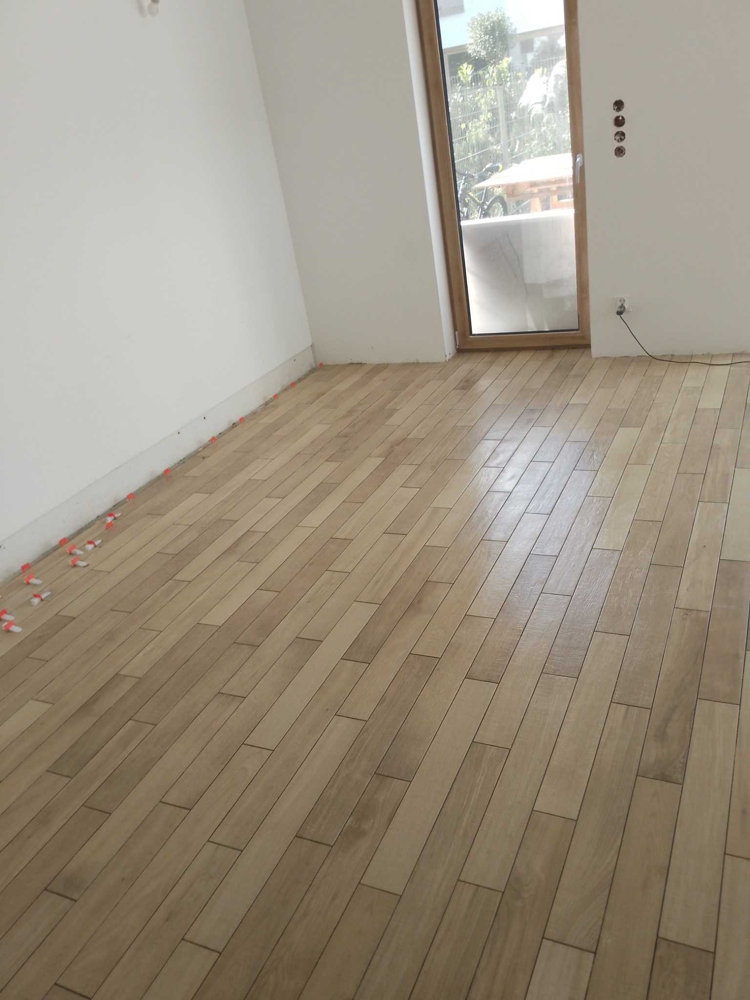 Układanie podłogi z paneli imitujących drewno w jasnym odcieniu, widoczne plastikowe krzyżyki dystansowe przy ścianie, surowy stan wnętrza z widocznymi gniazdkami elektrycznymi i oknem balkonowym.