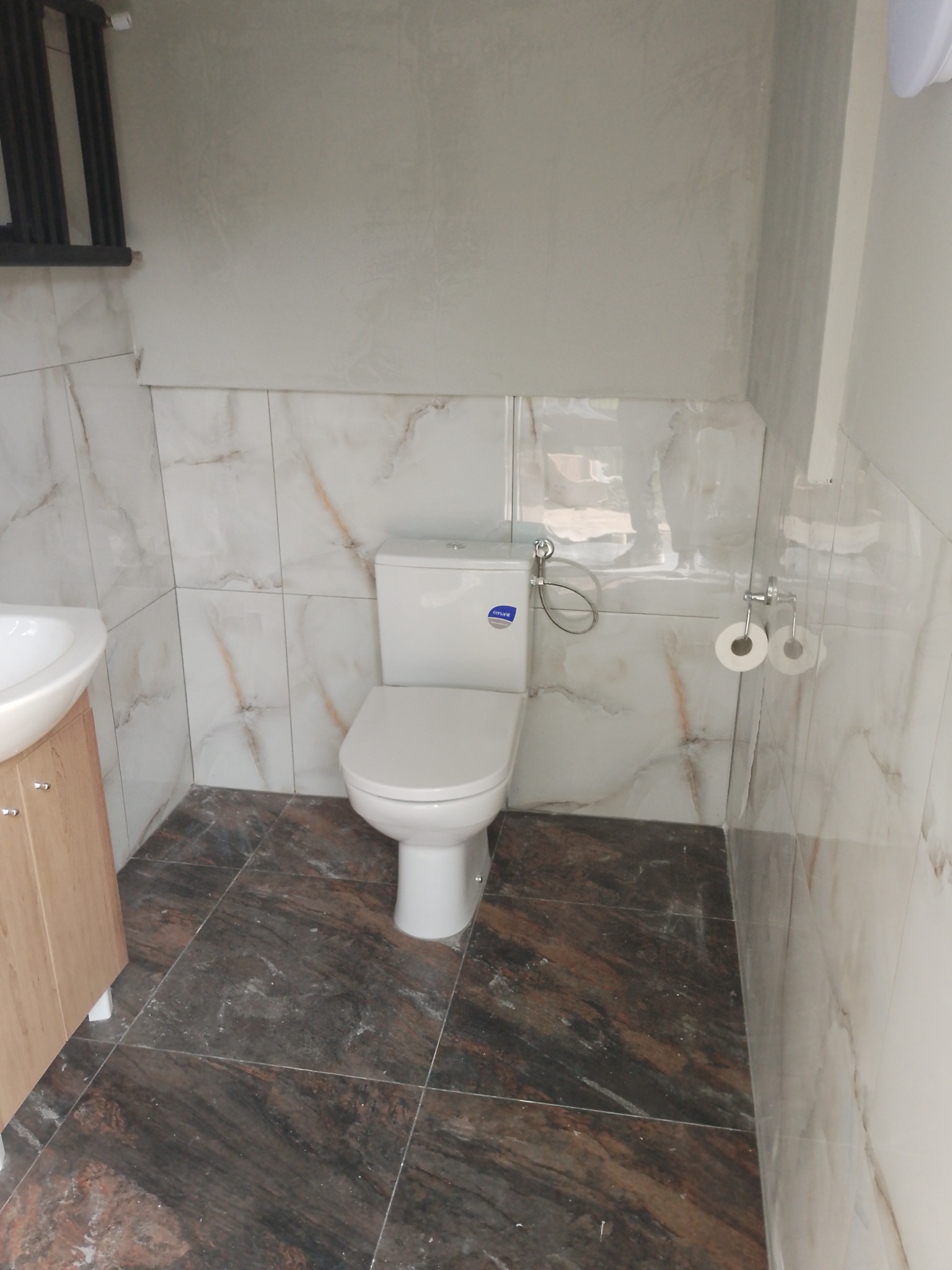 Wykończona łazienka z marmurowymi płytkami ściennymi i ciemnobrązowymi płytkami podłogowymi. Biała toaleta i umywalka z szafką. Widoczne akcesoria łazienkowe.