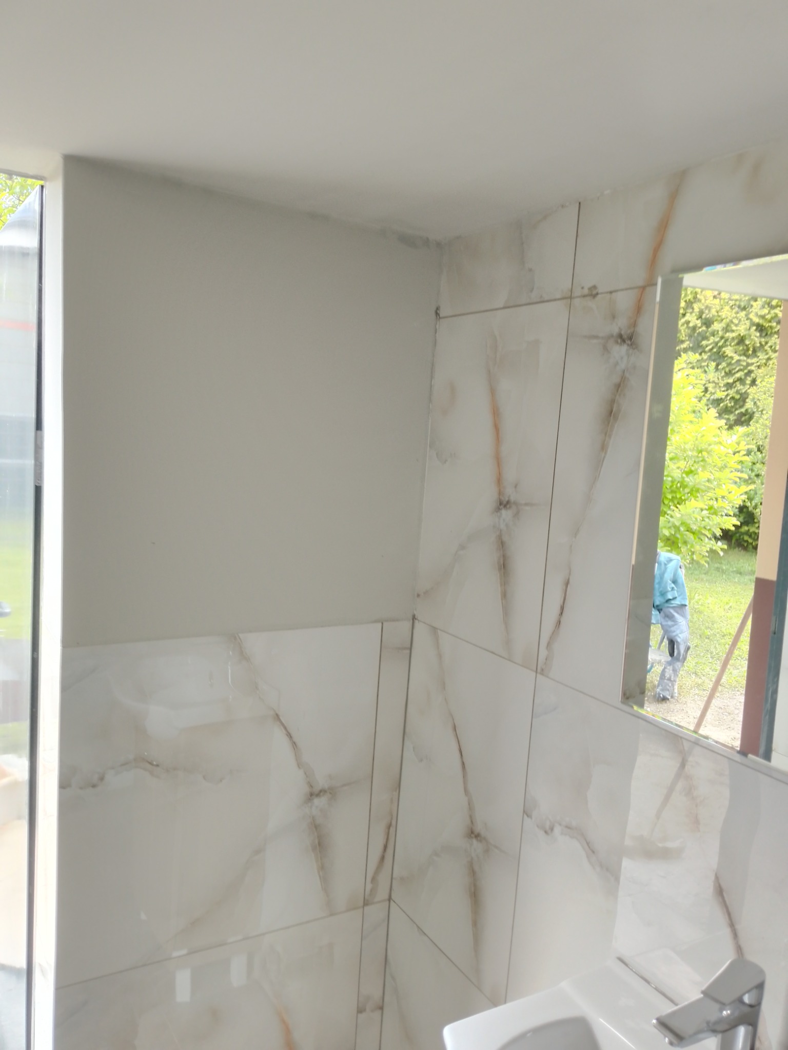 Ściany łazienki wyłożone białymi płytkami imitującymi marmur, z widocznym fragmentem umywalki i lustra, w którym odbija się postać pracownika na zewnątrz.