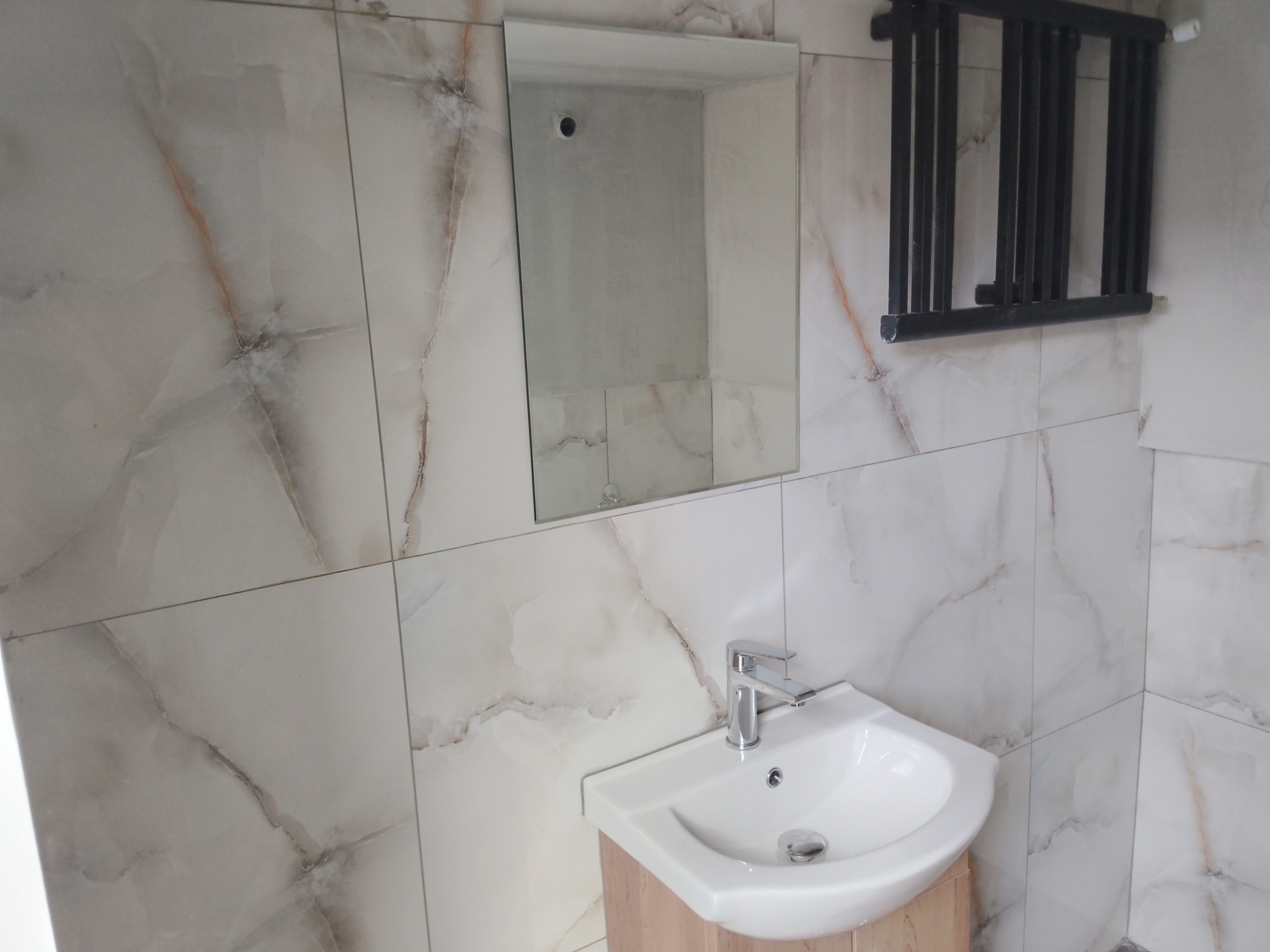 Wykończona łazienka z umywalką narożną, lustrem i grzejnikiem drabinkowym na ścianie z marmurowymi płytkami w odcieniach bieli i beżu. Nowoczesny design.