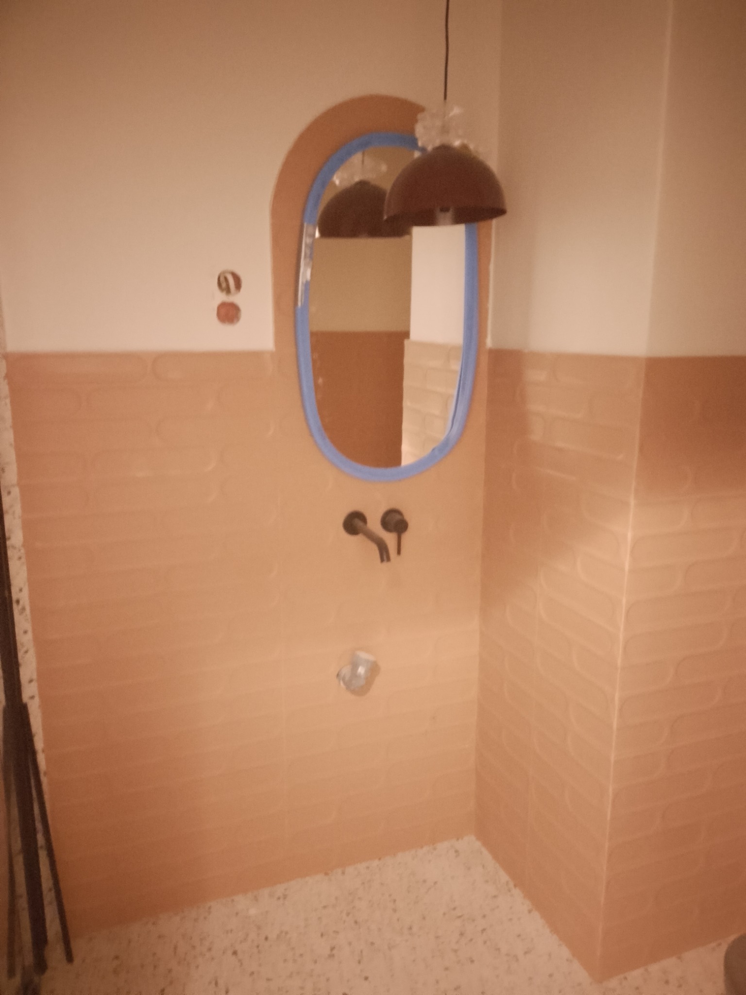 Wykończona łazienka z beżowymi płytkami o wzorze fal, owalnym lustrem z niebieską ramą i czarną armaturą. Widoczna lampa sufitowa i gniazdka elektryczne.