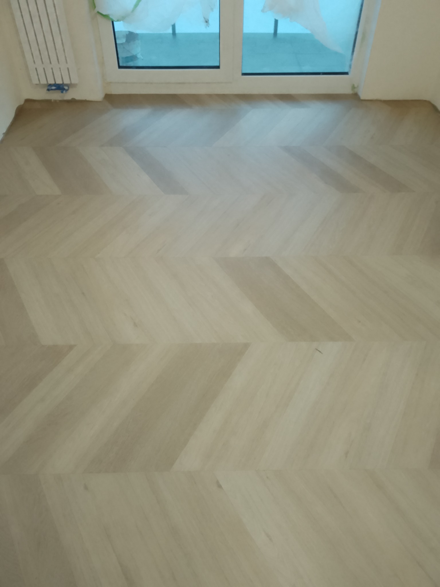 Jasne panele ułożone w jodełkę francuską, widok z góry. Grzejnik i okno w tle. Nowoczesne wykończenie podłogi w pokoju.