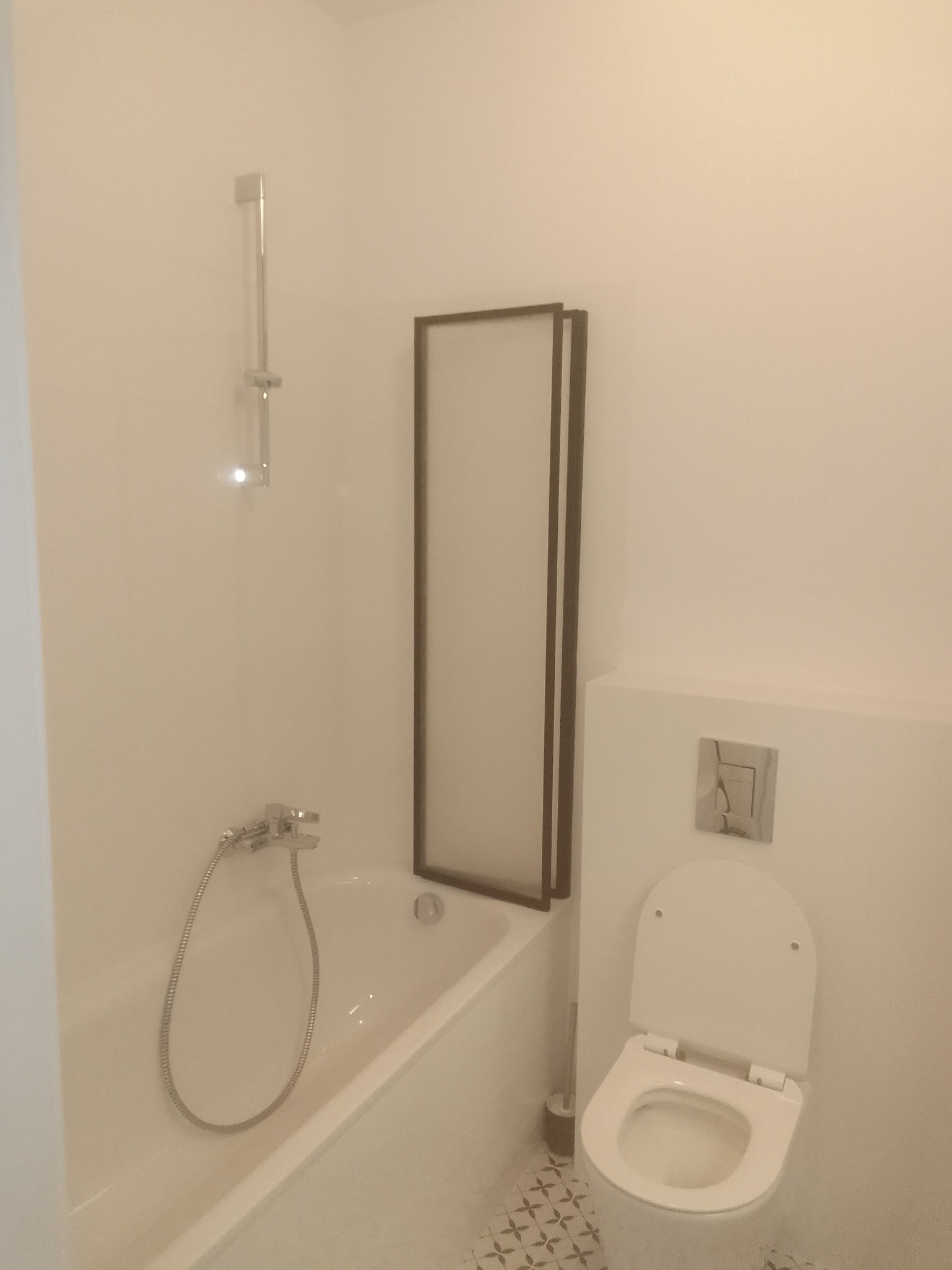 Jasna łazienka po remoncie: biała wanna z parawanem, toaleta z podtynkową spłuczką i wzorzysta podłoga. Minimalistyczny design.