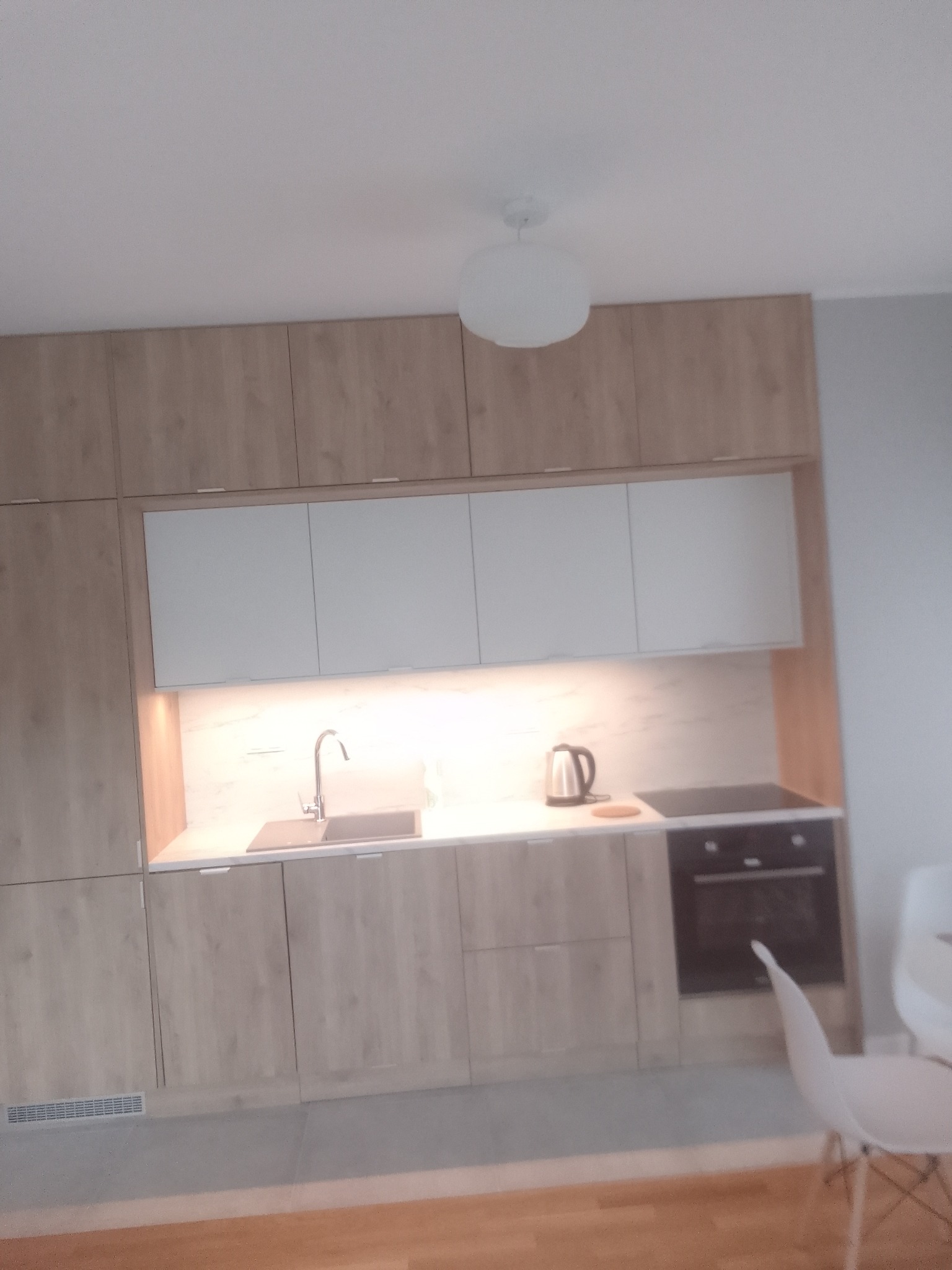 Jasna kuchnia z drewnianymi szafkami i białymi górnymi modułami. Widoczny zlew, płyta grzewcza i czajnik. Minimalistyczny wystrój wnętrza.