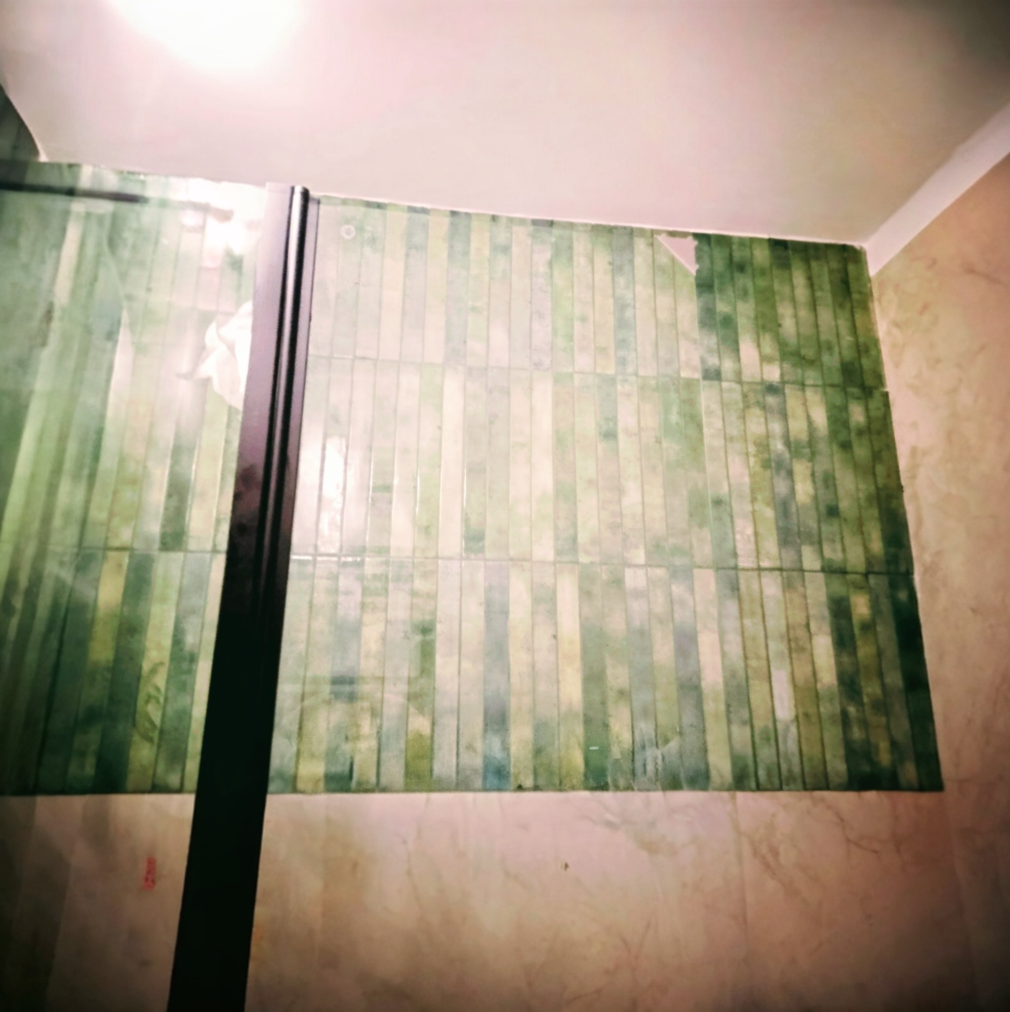 Ściana w łazience wyłożona wąskimi, pionowymi płytkami w odcieniach zieleni i beżu, widoczna kabina prysznicowa z czarną ramą. Górna część ściany bez płytek.