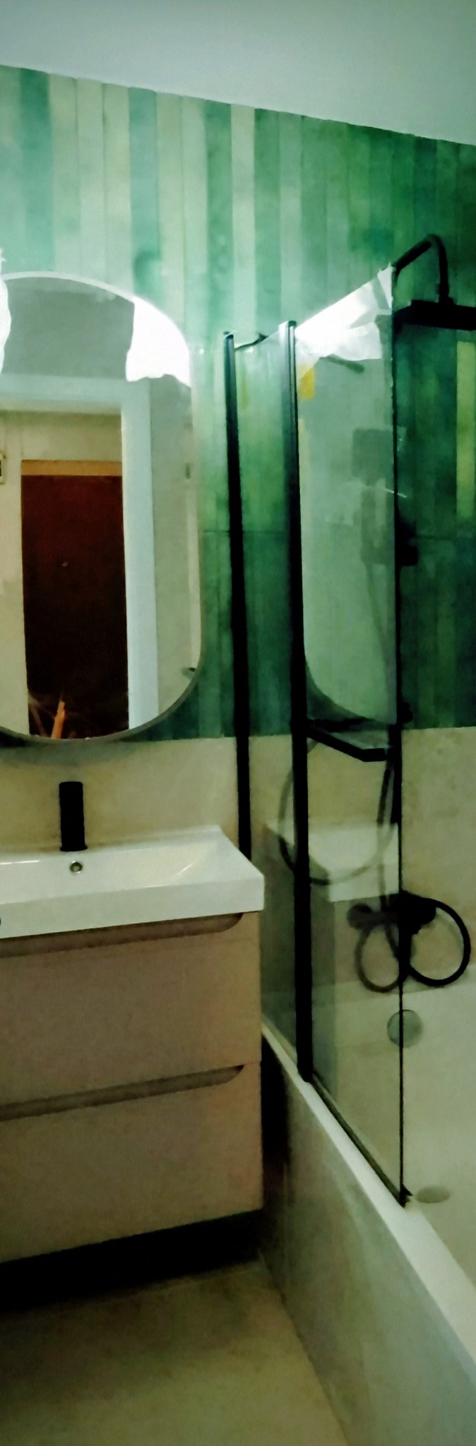 Nowoczesna łazienka z kabiną prysznicową typu walk-in, umywalką z szafką i owalnym lustrem. Ściany w pionowe pasy zieleni i beżu. Minimalistyczny design.