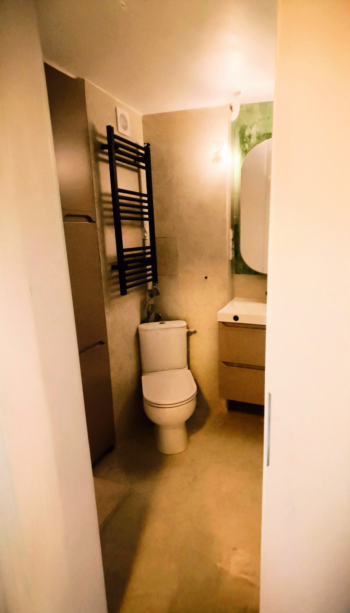 Wykończona łazienka w beżach: toaleta, grzejnik drabinkowy, szafka z umywalką. Minimalistyczny design, ciepłe oświetlenie, widok zza uchylonych drzwi.