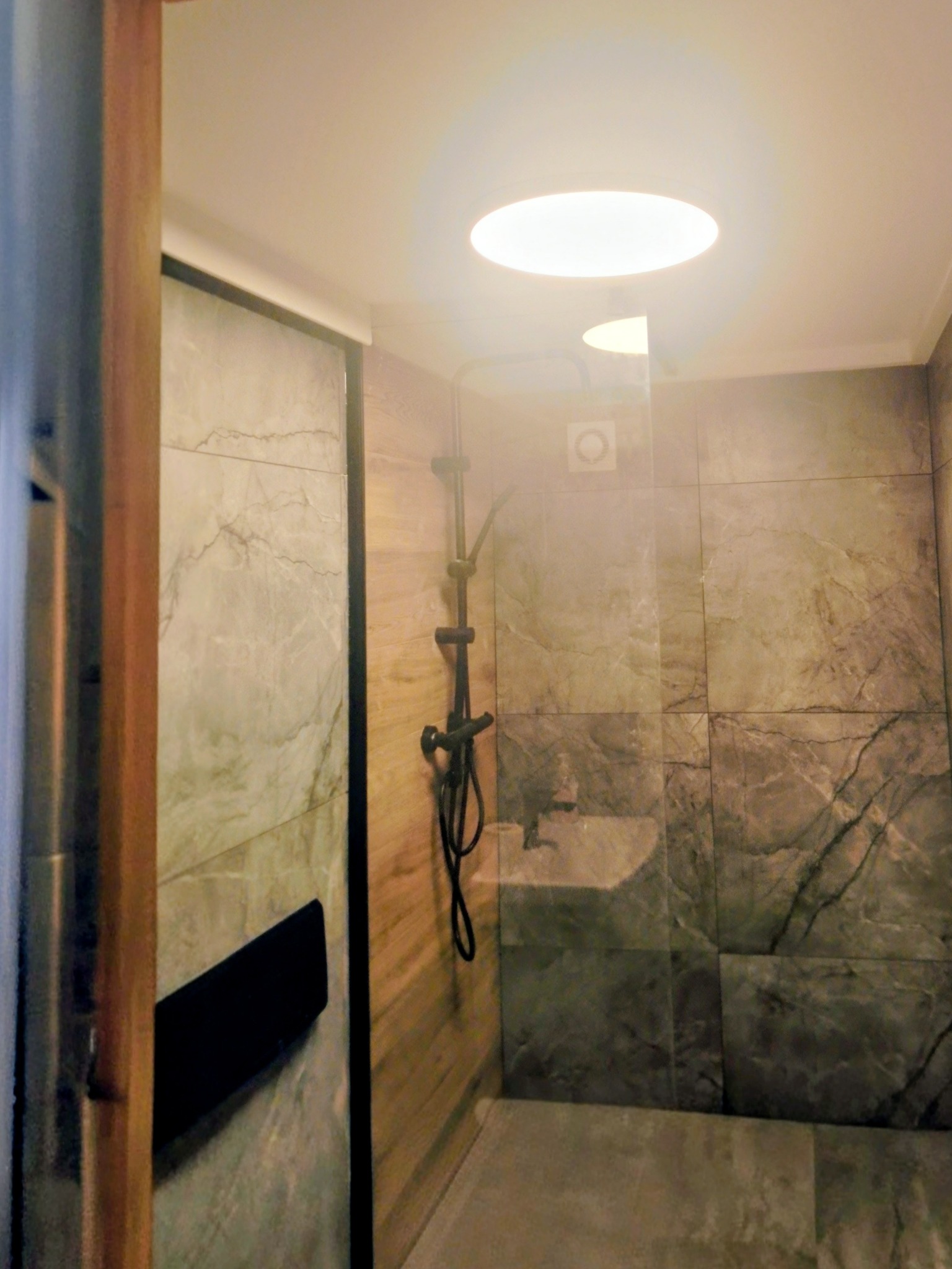 Nowoczesna kabina prysznicowa z szarymi płytkami imitującymi marmur, czarną armaturą i oświetleniem punktowym. Widoczny fragment drewnianej ściany.