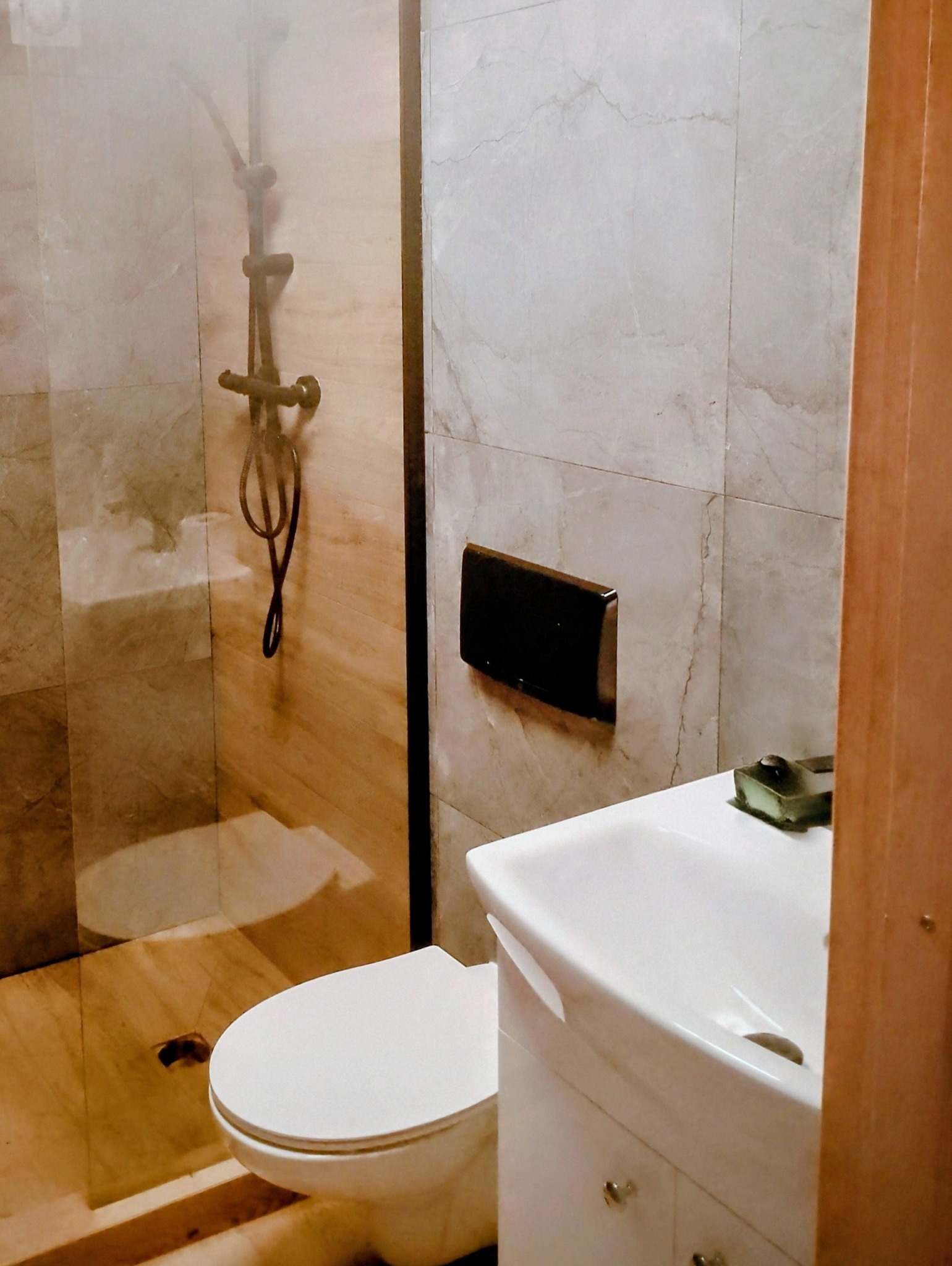 Nowoczesna łazienka z marmurowymi płytkami, kabiną prysznicową typu walk-in i białą umywalką z szafką. Widoczny czarny przycisk spłuczki i armatura w kolorze ciemnego brązu.