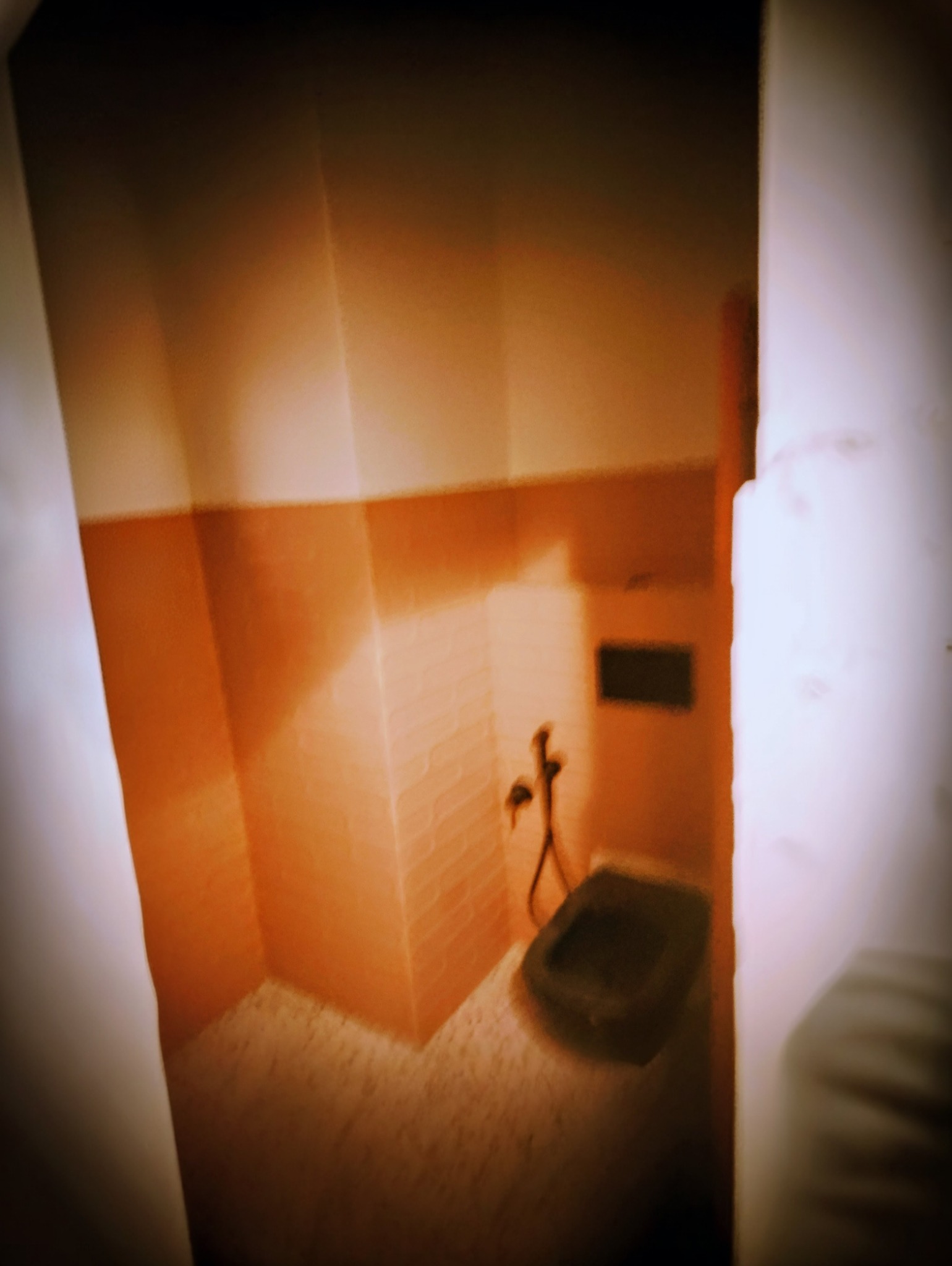 Narożna umywalka w łazience wykończonej pomarańczowymi płytkami, widok zza framugi drzwi. Ujęcie z bliska, ciemne oświetlenie.