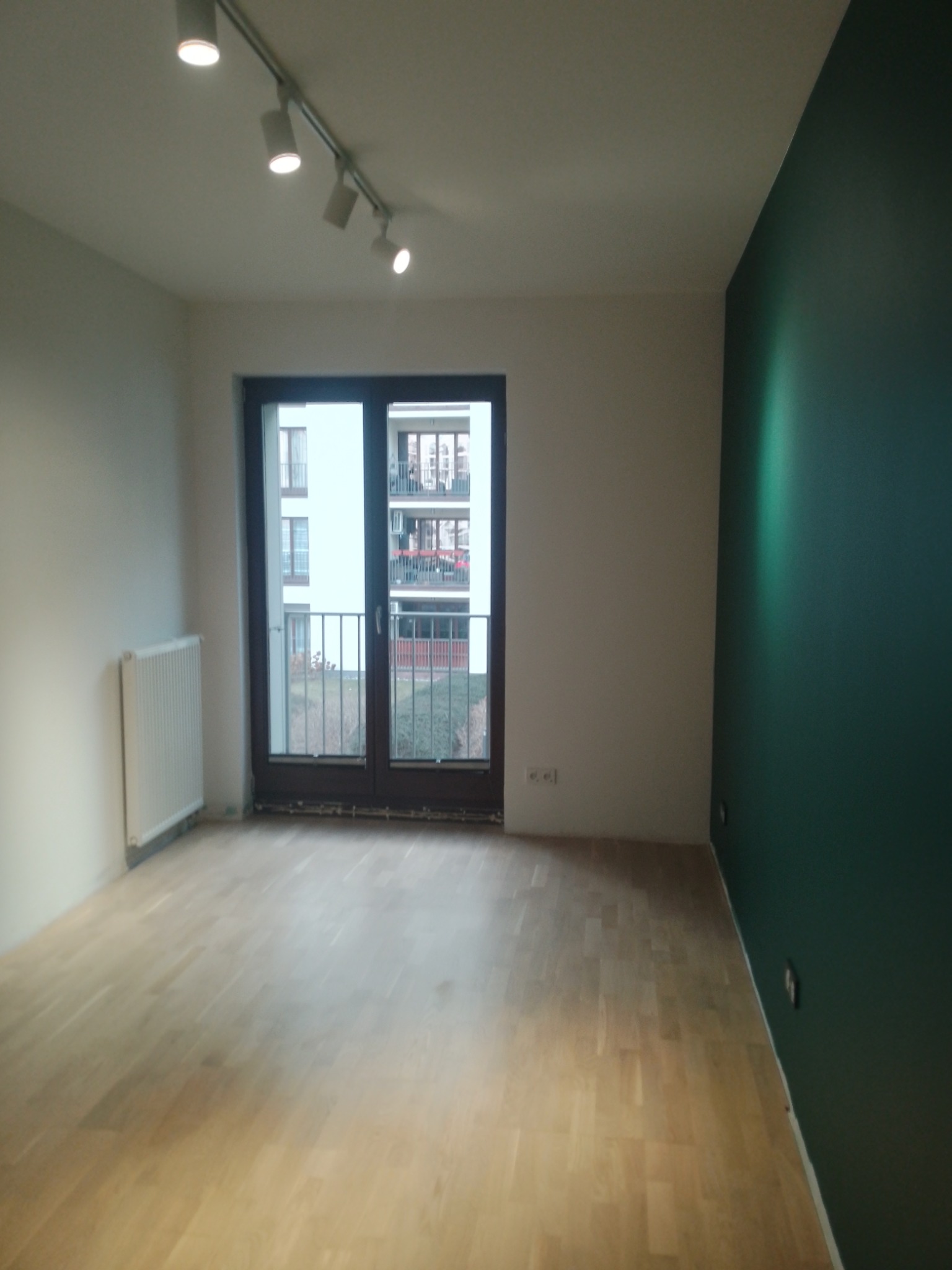 Jasne panele podłogowe w pokoju z ciemnozieloną ścianą i dużym oknem balkonowym. Widoczny grzejnik i oświetlenie sufitowe.