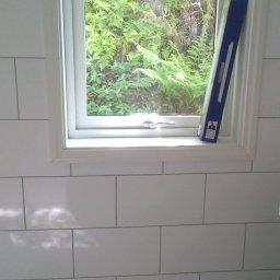 Prywatny wykonawca - Białe, prostokątne płytki ścienne z widocznymi krzyżykami dystansowymi u dołu, okno z widokiem na zieleń i poziomica oparta o ramę okienną.