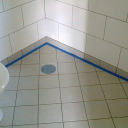 Prywatny wykonawca - Wykończona kabina prysznicowa z białymi płytkami ściennymi i podłogowymi, zabezpieczona niebieską taśmą malarską wzdłuż krawędzi, z widocznym odpływem liniowym i toaletą po lewej stronie.