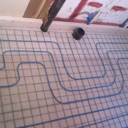 Prywatny wykonawca - Przygotowanie do wylewki pod ogrzewanie podłogowe: rury grzewcze w kolorze niebieskim ułożone na siatce stalowej, widoczny odpływ liniowy w rogu pomieszczenia i fragment instalacji kanalizacyjnej...