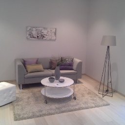 Prywatny wykonawca - Jasny salon z szarą sofą, stolikiem kawowym na kółkach i lampą stojącą, doświetlony światłem z okna z widokiem na kamienny mur.