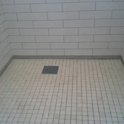 Wykończony brodzik z białymi, prostokątnymi płytkami na ścianach i kwadratowymi na podłodze, z szarą listwą przypodłogową i odpływem liniowym.