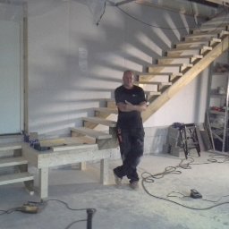 Prywatny wykonawca - Stolarz oparty o surowe drewniane schody w trakcie budowy, widoczne narzędzia, nieotynkowana ściana i prowizoryczne oświetlenie.