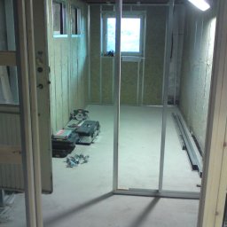 Prywatny wykonawca - Wnętrze w trakcie prac wykończeniowych, widoczne metalowe profile ścian działowych, ocieplenie wełną mineralną, narzędzia i skrzynki narzędziowe na podłodze, okno z widokiem na zewnątrz.