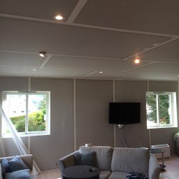 Prywatny wykonawca - Pokój w trakcie remontu: ściany z płyt gipsowo-kartonowych, widoczne profile na suficie, dwa okna z widokiem na zieleń, telewizor na ścianie, częściowo umeblowany z sofą i stolikiem kawowym, taśma...