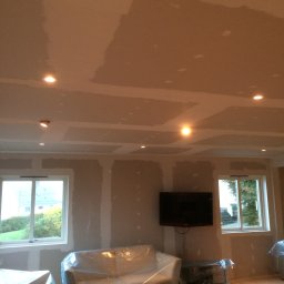 Prywatny wykonawca - Pokój w trakcie prac przygotowawczych: ściany i sufit po nałożeniu gładzi, zabezpieczone meble folią, widoczne okna z widokiem na zieleń i zamontowany telewizor.