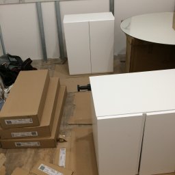 Prywatny wykonawca - Białe szafki w trakcie montażu, widoczne kartony z oznaczeniem 'KUCHNIA ASPEN 2M' oraz elementy montażowe na podłodze, metalowe profile na ścianie w tle.