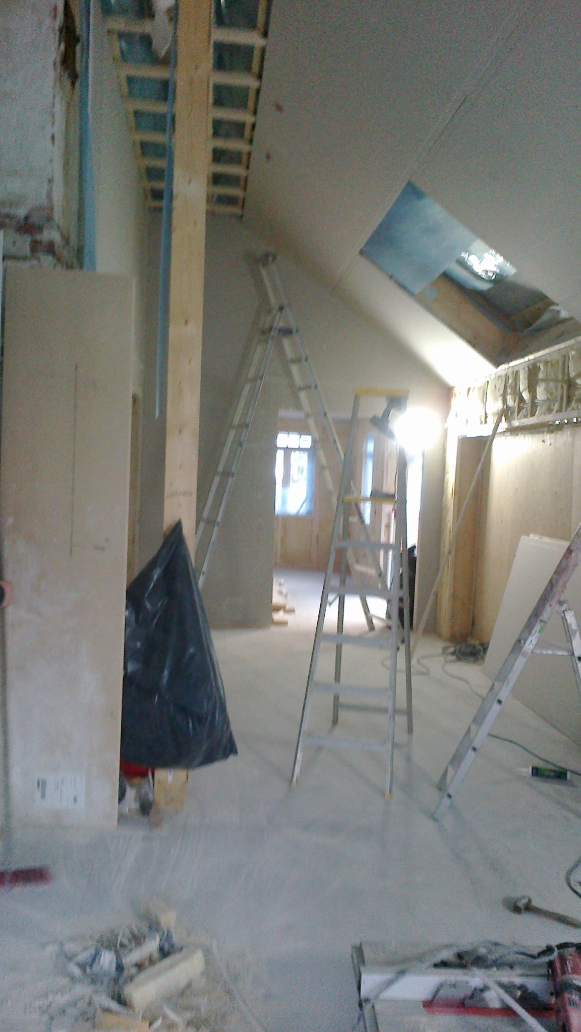 Wnętrze w trakcie prac wykończeniowych: widoczne dwie drabiny, surowe ściany, przygotowane płyty kartonowo-gipsowe, otwór okienny w dachu i drewniana konstrukcja pod sufitem.