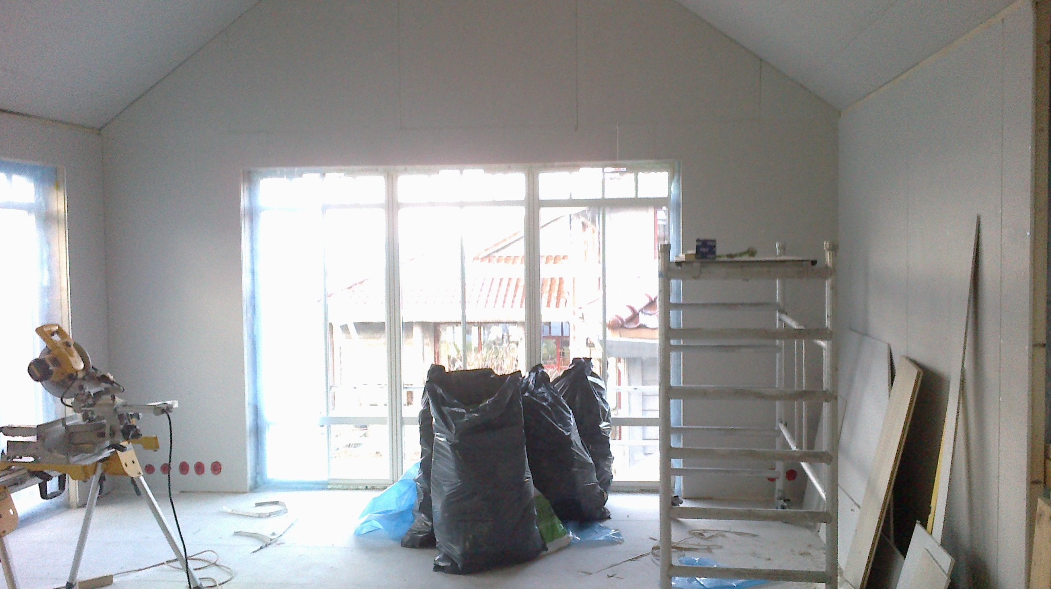 Wnętrze w trakcie prac: widoczna piła ukosowa na statywie, worki z odpadami budowlanymi, aluminiowy ruszt i płyty kartonowo-gipsowe oparte o ścianę, okno zabezpieczone folią.