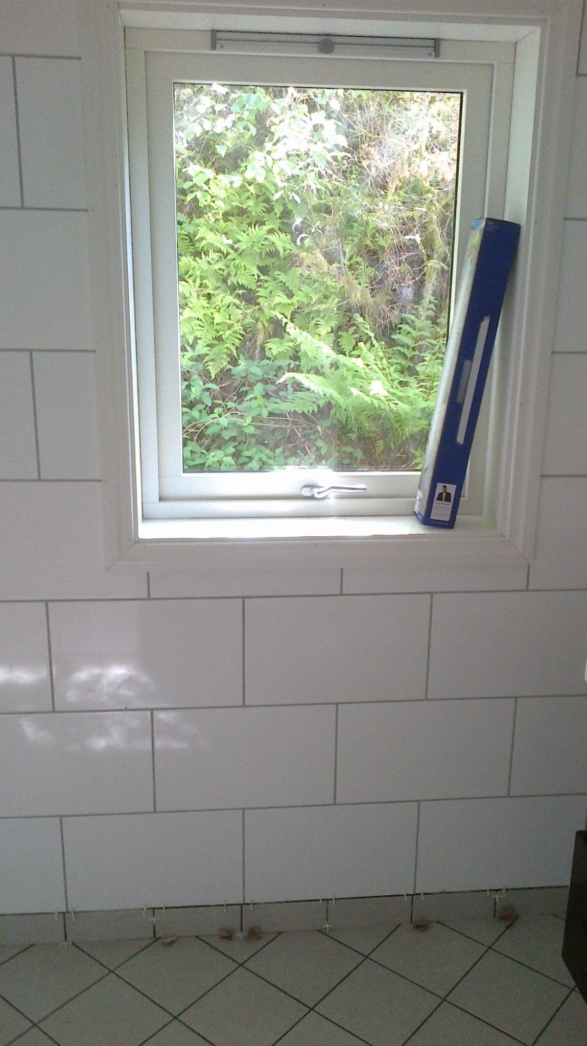 Białe, prostokątne płytki ścienne z widocznymi krzyżykami dystansowymi u dołu, okno z widokiem na zieleń i poziomica oparta o ramę okienną.