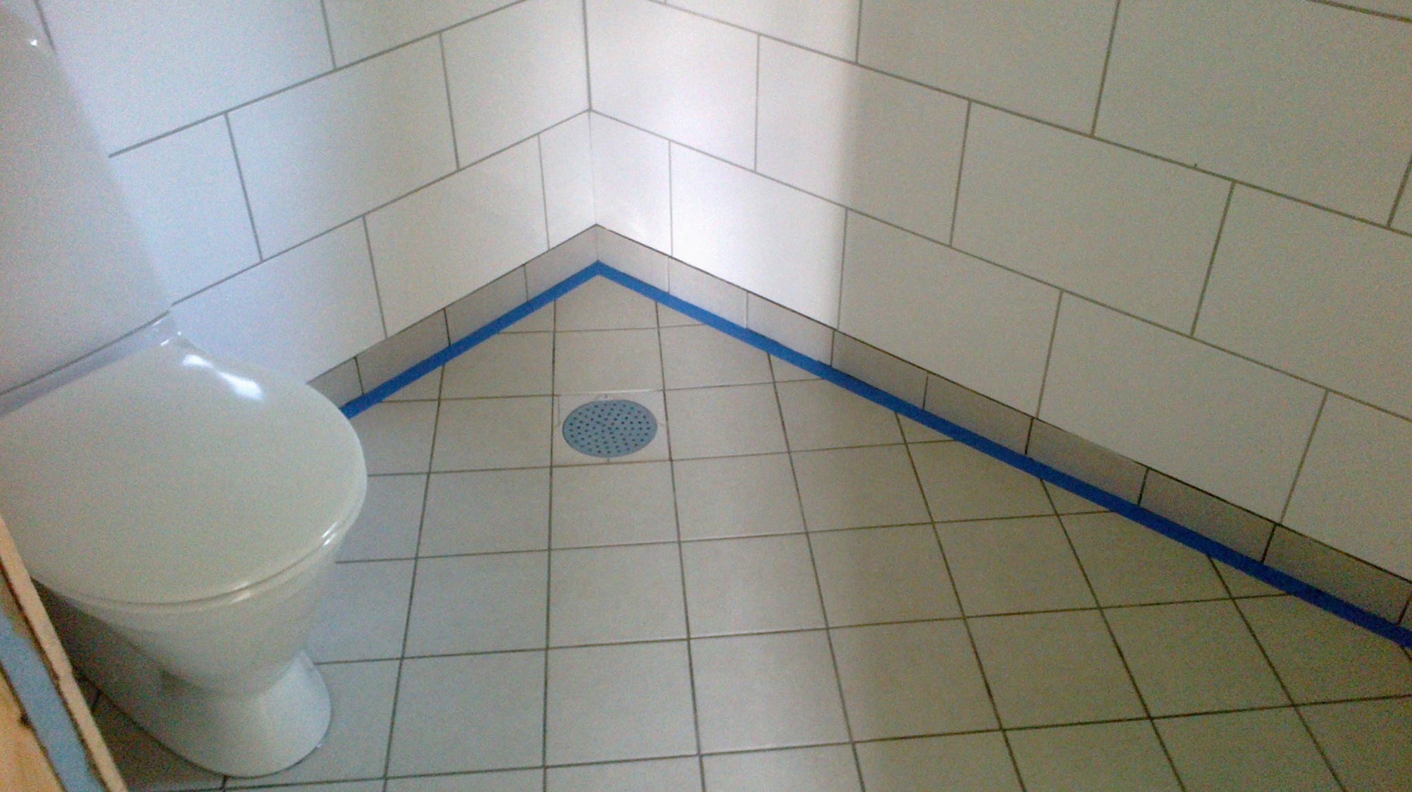 Wykończona kabina prysznicowa z białymi płytkami ściennymi i podłogowymi, zabezpieczona niebieską taśmą malarską wzdłuż krawędzi, z widocznym odpływem liniowym i toaletą po lewej stronie.