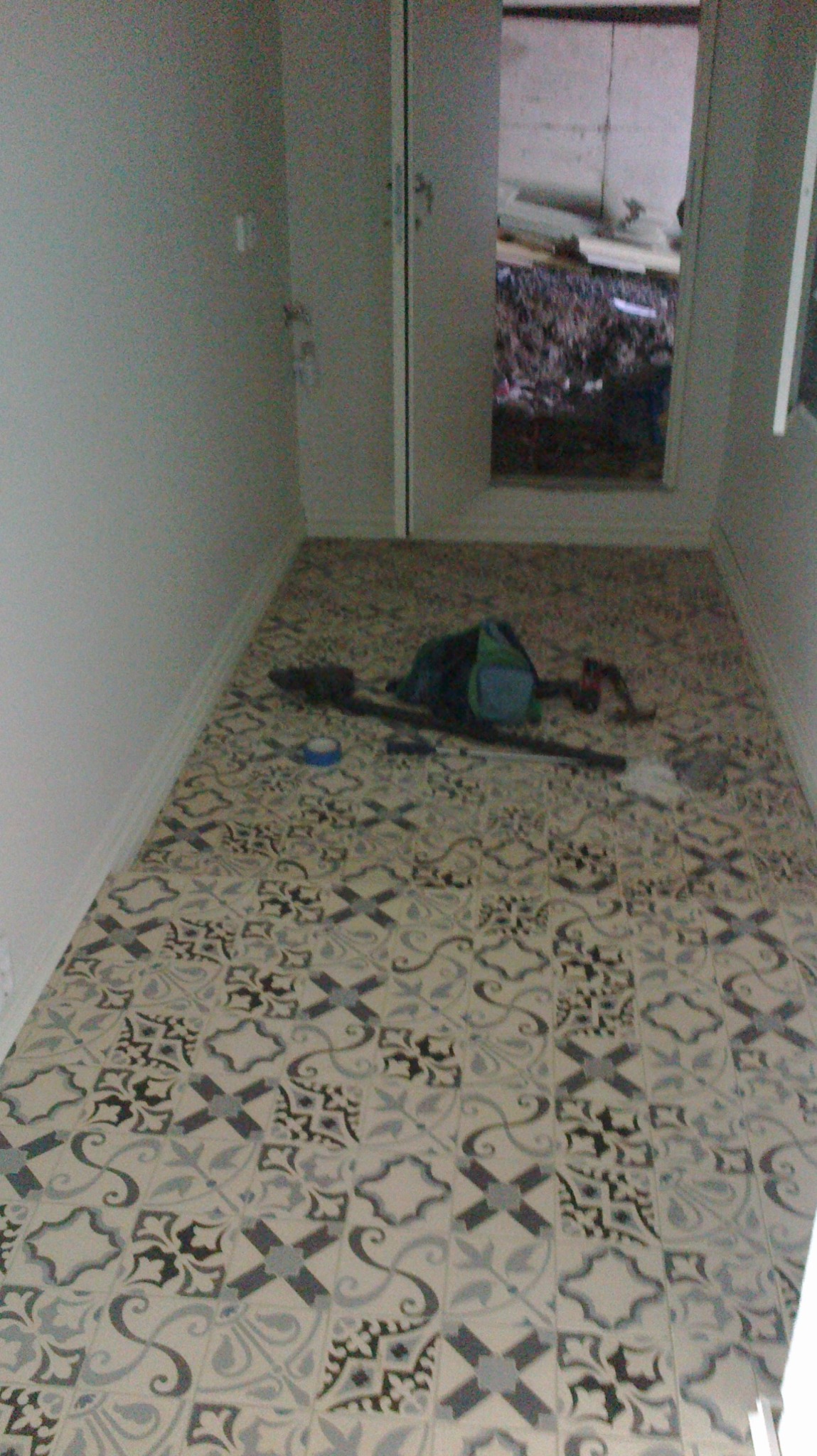 Wnętrze korytarza z nowo ułożoną, wzorzystą podłogą z płytek ceramicznych w odcieniach szarości i beżu, widoczne narzędzia glazurnicze i torba majsterkowicza, otwarte drzwi do pomieszczenia...