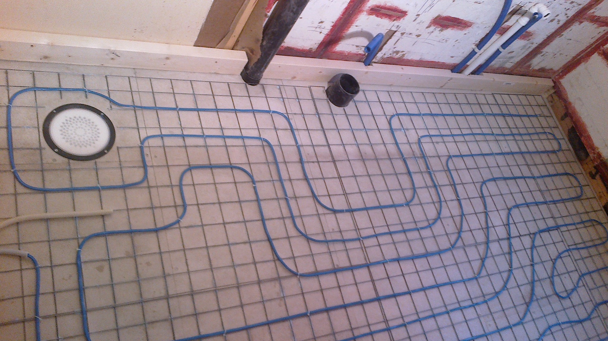 Przygotowanie do wylewki pod ogrzewanie podłogowe: rury grzewcze w kolorze niebieskim ułożone na siatce stalowej, widoczny odpływ liniowy w rogu pomieszczenia i fragment instalacji kanalizacyjnej...