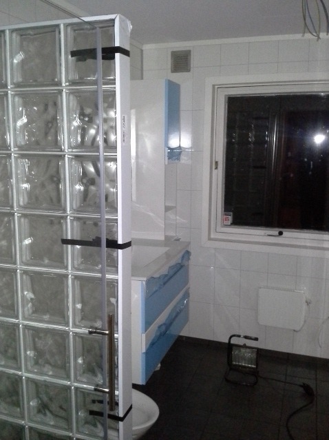 Łazienka w trakcie prac: ściana z luksferów, biało-błękitna szafka pod umywalkę z folią ochronną, okno, czarna podłoga i lampa robocza.