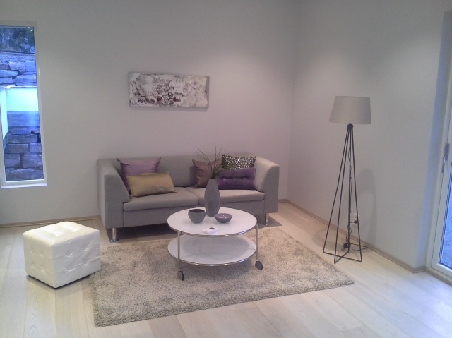 Jasny salon z szarą sofą, stolikiem kawowym na kółkach i lampą stojącą, doświetlony światłem z okna z widokiem na kamienny mur.