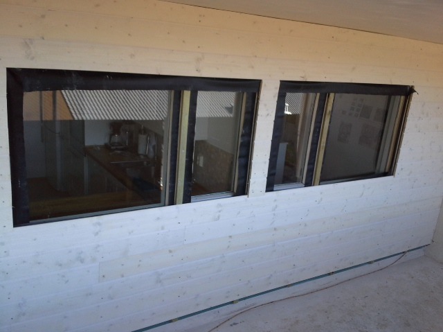 Wnętrze w trakcie prac wykończeniowych: trzy okna z czarną taśmą uszczelniającą osadzone w ścianie z jasnych, drewnianych paneli boazeryjnych.