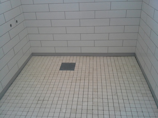 Wykończony brodzik z białymi, prostokątnymi płytkami na ścianach i kwadratowymi na podłodze, z szarą listwą przypodłogową i odpływem liniowym.