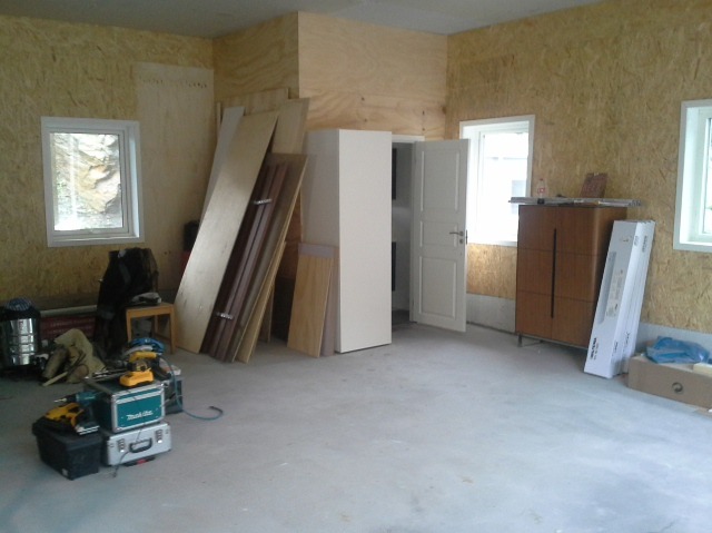 Wnętrze w trakcie remontu: ściany wyłożone płytami OSB, okna z widokiem na zewnątrz, drzwi, meble, narzędzia i płyty czekające na montaż.