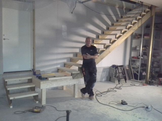 Stolarz oparty o surowe drewniane schody w trakcie budowy, widoczne narzędzia, nieotynkowana ściana i prowizoryczne oświetlenie.