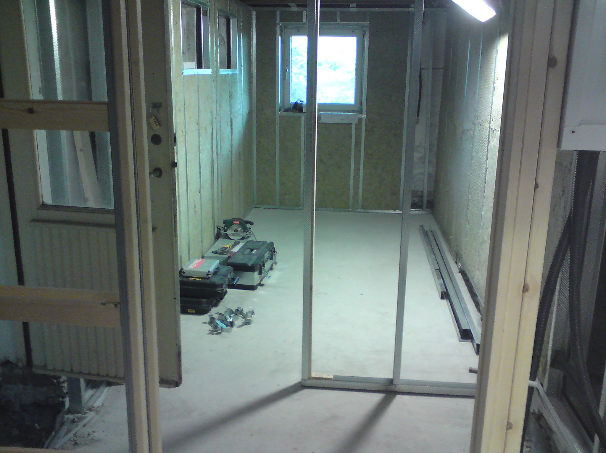 Wnętrze w trakcie prac wykończeniowych, widoczne metalowe profile ścian działowych, ocieplenie wełną mineralną, narzędzia i skrzynki narzędziowe na podłodze, okno z widokiem na zewnątrz.