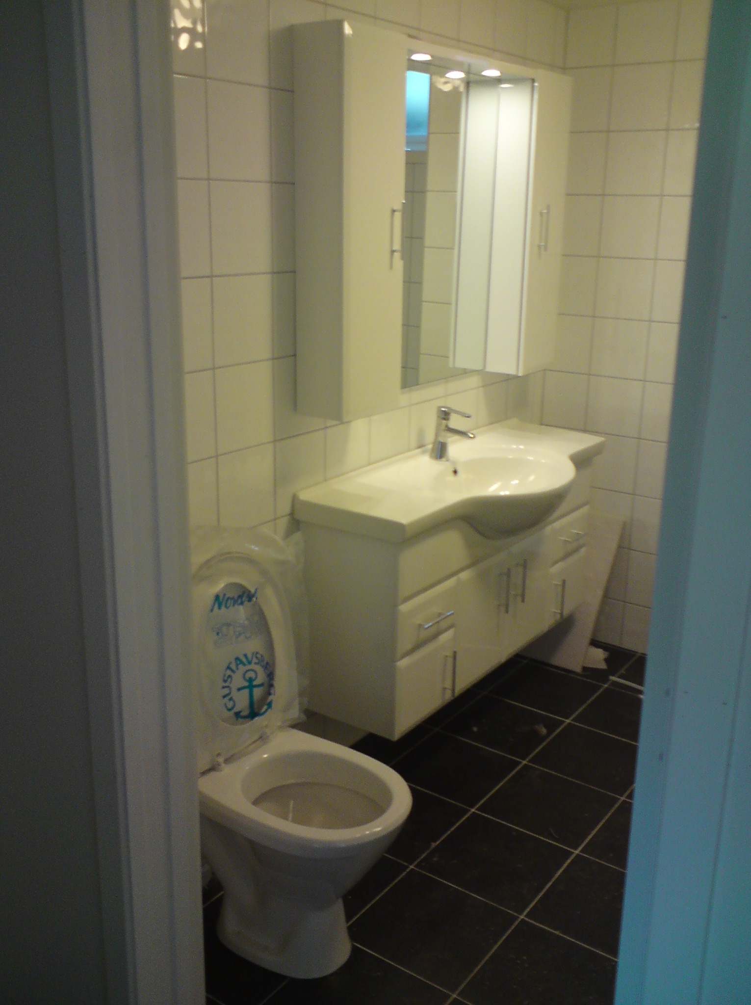 Biała łazienka z czarną podłogą, widok na umywalkę z szafką i lustrem oraz toaletę z deską z napisem 'Gustavsberg'.