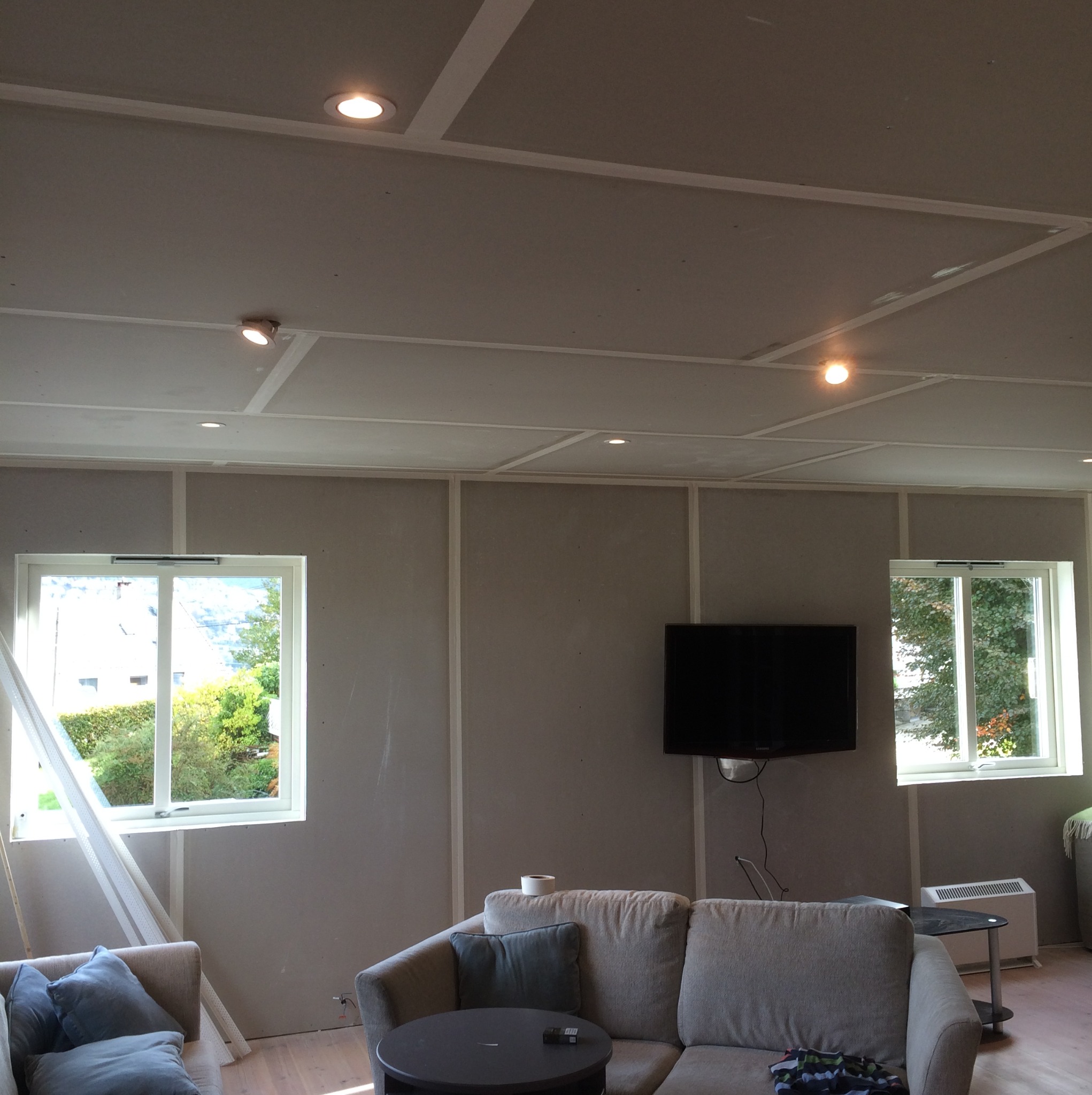 Pokój w trakcie remontu: ściany z płyt gipsowo-kartonowych, widoczne profile na suficie, dwa okna z widokiem na zieleń, telewizor na ścianie, częściowo umeblowany z sofą i stolikiem kawowym, taśma...