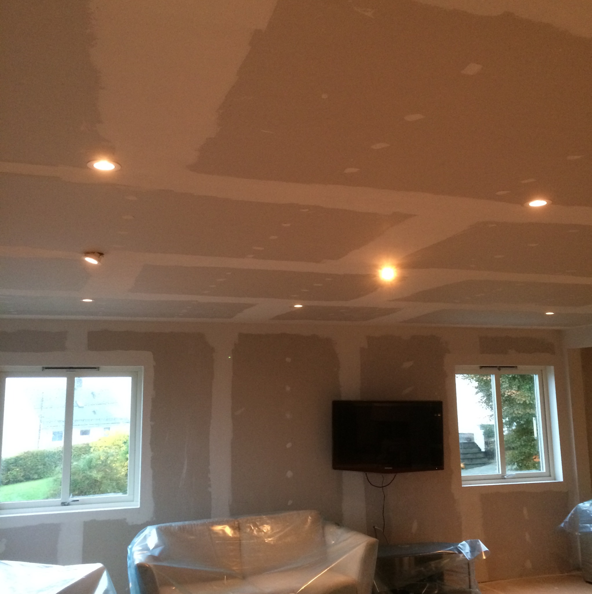 Pokój w trakcie prac przygotowawczych: ściany i sufit po nałożeniu gładzi, zabezpieczone meble folią, widoczne okna z widokiem na zieleń i zamontowany telewizor.