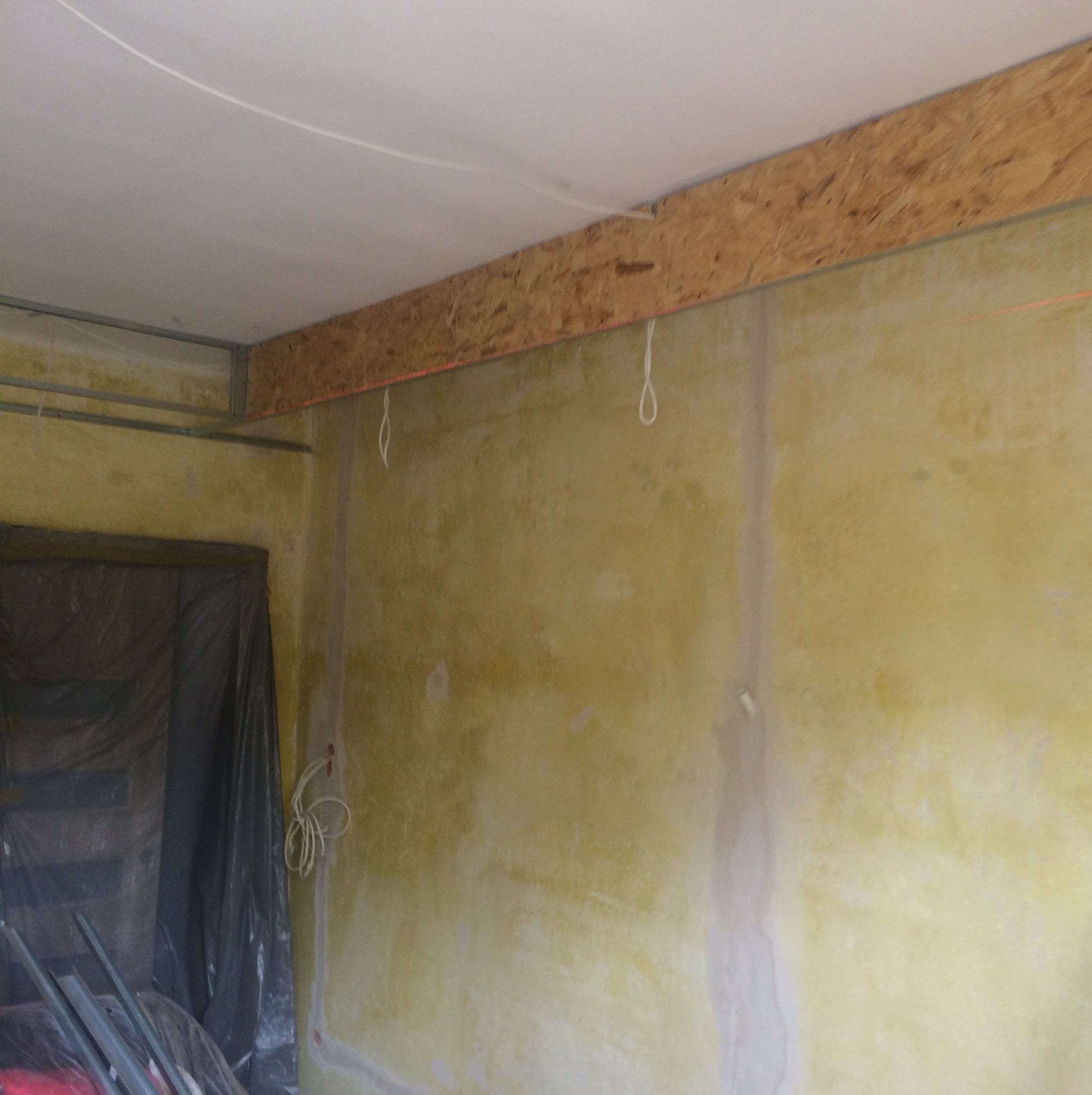 Ściana w trakcie przygotowania do malowania, z widoczną warstwą podkładową, drewnianą belką pod sufitem i folią ochronną na podłodze.