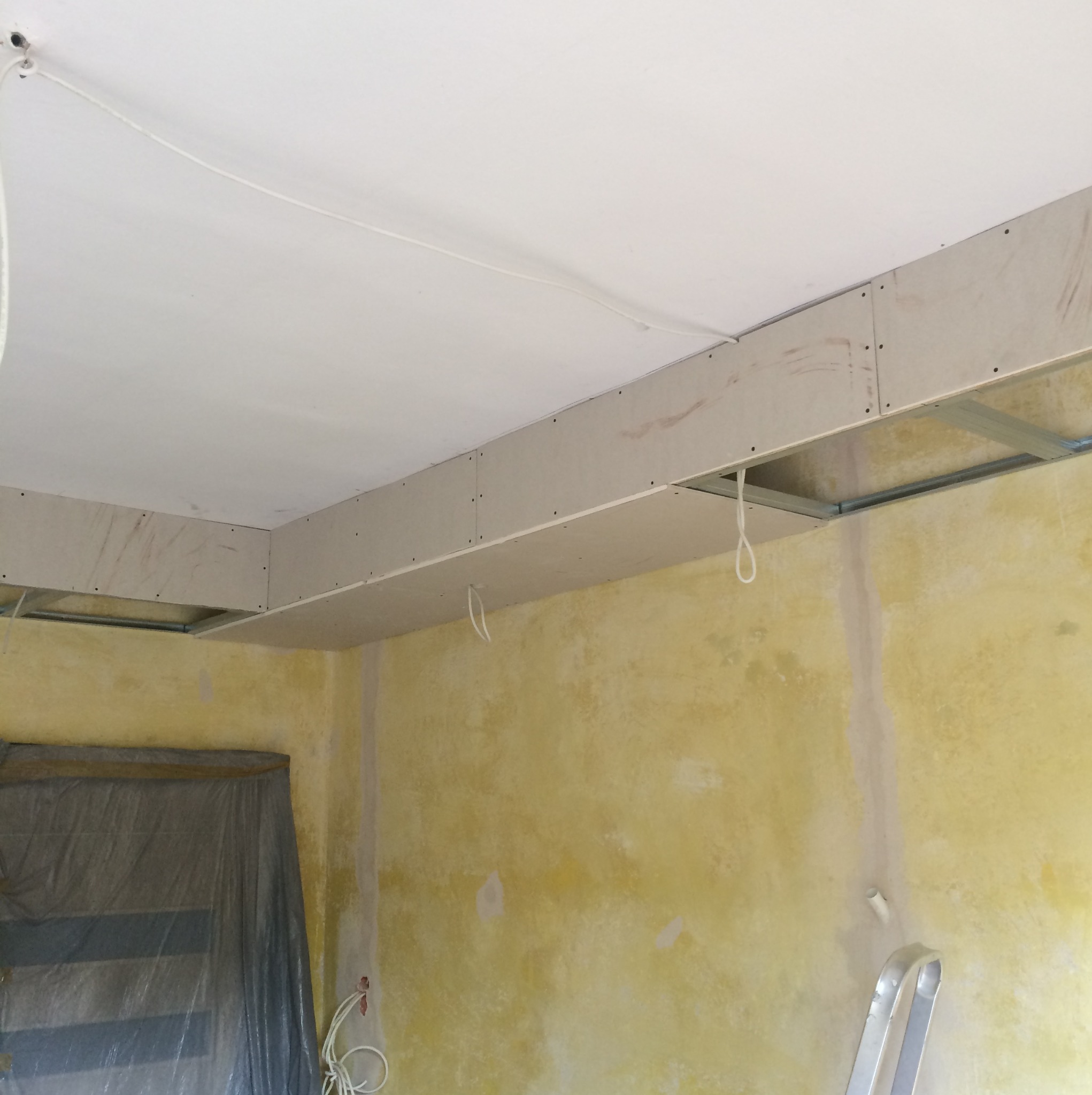 Sufit podwieszany z płyt gipsowo-kartonowych na metalowej konstrukcji, widoczne przewody elektryczne, ściany w żółtym kolorze przed malowaniem, zabezpieczone okno folią malarską.