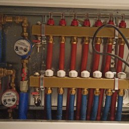 Tomasz Studziński Instalacje - Rozdzielacz ogrzewania podłogowego w metalowej szafce z rurami w kolorach niebieskim i czerwonym, z wodomierzami i licznikiem ciepła SanTech.