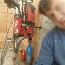 Tomasz Studziński Instalacje - Pracownik wierci otwór w ceglanej ścianie za pomocą wiertnicy rdzeniowej. Widoczny fragment twarzy pracownika i narzędzie pracy.
