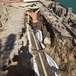 Tomasz Studziński Instalacje - Instalacja rur w wykopie, widok z góry. Mężczyzna w wykopie układa rury na geowłókninie. Na górze kostka brukowa i gruz. Praca w toku.