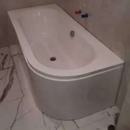 Nowoczesna, biała wanna narożna z chromowanym odpływem, zamontowana w łazience z marmurowymi płytkami. Widoczne narzędzia hydrauliczne na podłodze.