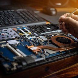 Tecza - Otwarty laptop z widoczną płytą główną, wentylatorem i elementami elektronicznymi, ręka trzyma precyzyjny śrubokręt, klawiatura w tle, ciepłe oświetlenie.