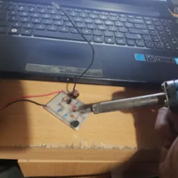 Lutowanie elementów elektronicznych na płytce drukowanej, widoczny laptop w tle, zasilanie z baterii 9V.