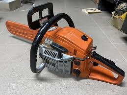 Pomarańczowa piła spalinowa leżąca na szarej, kwadratowej podłodze z płytek. Widoczna czarna rękojeść i metalowa obudowa silnika. W tle fragmenty innych narzędzi.